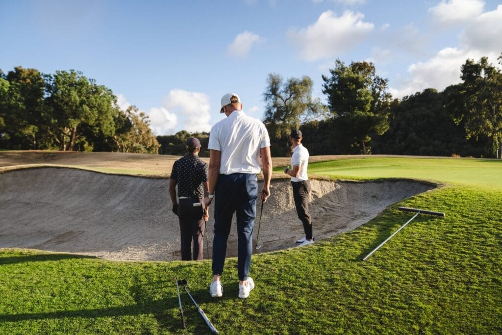 Aaron Rai, on the left in a dark shirt, receives coaching on bunker shots from his two PGA Tour coaches, Piers Ward and Andy Proudman, during a golf training session.